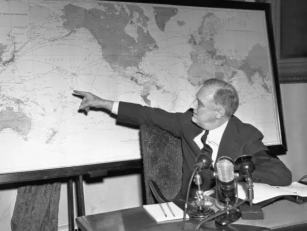 President Roosevelt at a desk with microphones in front of him, pointing at a wall map.